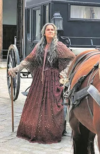 A atriz Ingrid Guimarães com longos cabelos brancos e um vestido vinho como a Elvira com um cavalo a sua frente em cena de Nos Tempos do Imperador