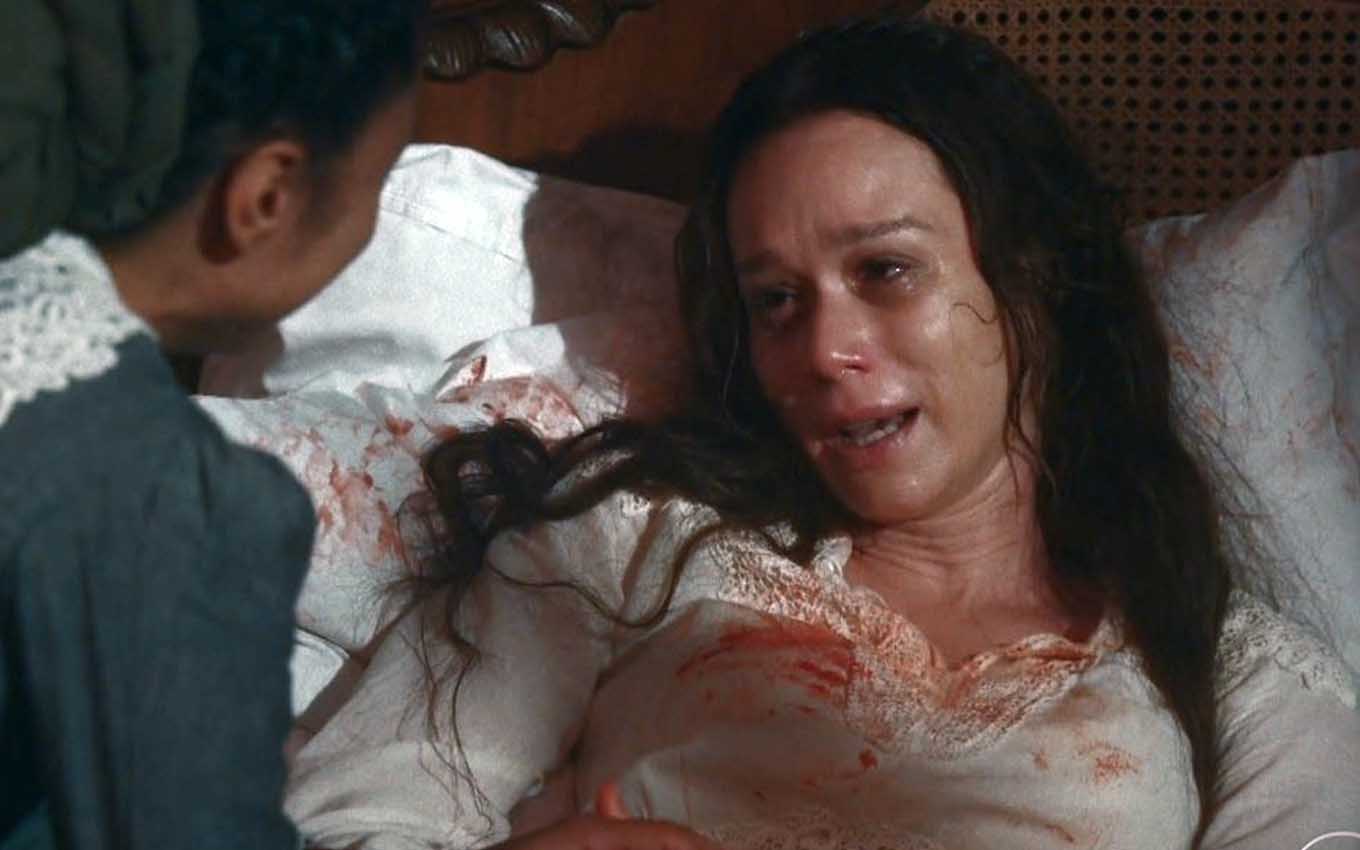 A atriz Mariana Ximenes está em uma cama, com olheiras e aparência péssima, com a roupa manchada de sangue, como a Luísa em cena de Nos Tempos do Imperador