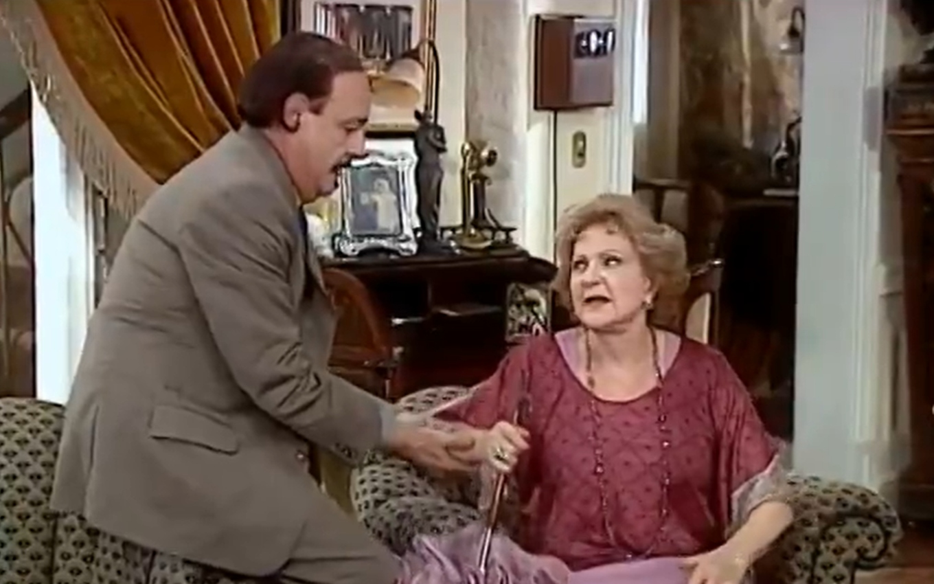 Ney Latorraca, caracterizado como Cornélio, ajuda Eva Todor, a Josefa, se sentar no sofá em cena de O Cravo e a Rosa