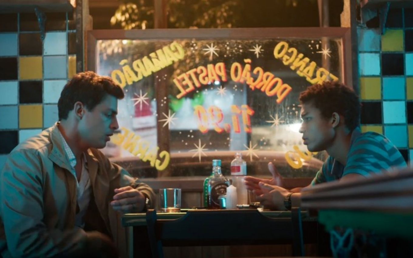 Do lado esquerdo de uma mesa de bar, o ator Cauã Reymond como Christian olha para Juan Paiva, o Ravi, do lado esquerdo em cena noturna de Um Lugar ao Sol