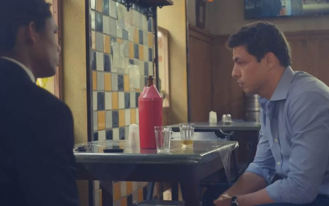 O ator Juan Paiva, de costas, como Ravi bebe cerveja com Cauã Reymond, o Christian, em um bar em cena de Um Lugar ao Sol
