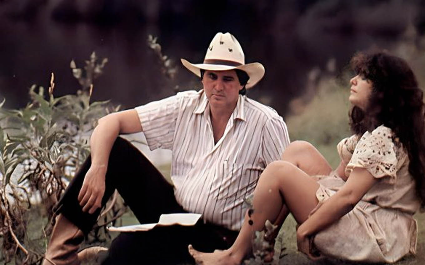 O cantor Sérgio Reis como Tibério ao lado de Andréa Richa, a Muda, sentados em um campo em cena de Pantanal