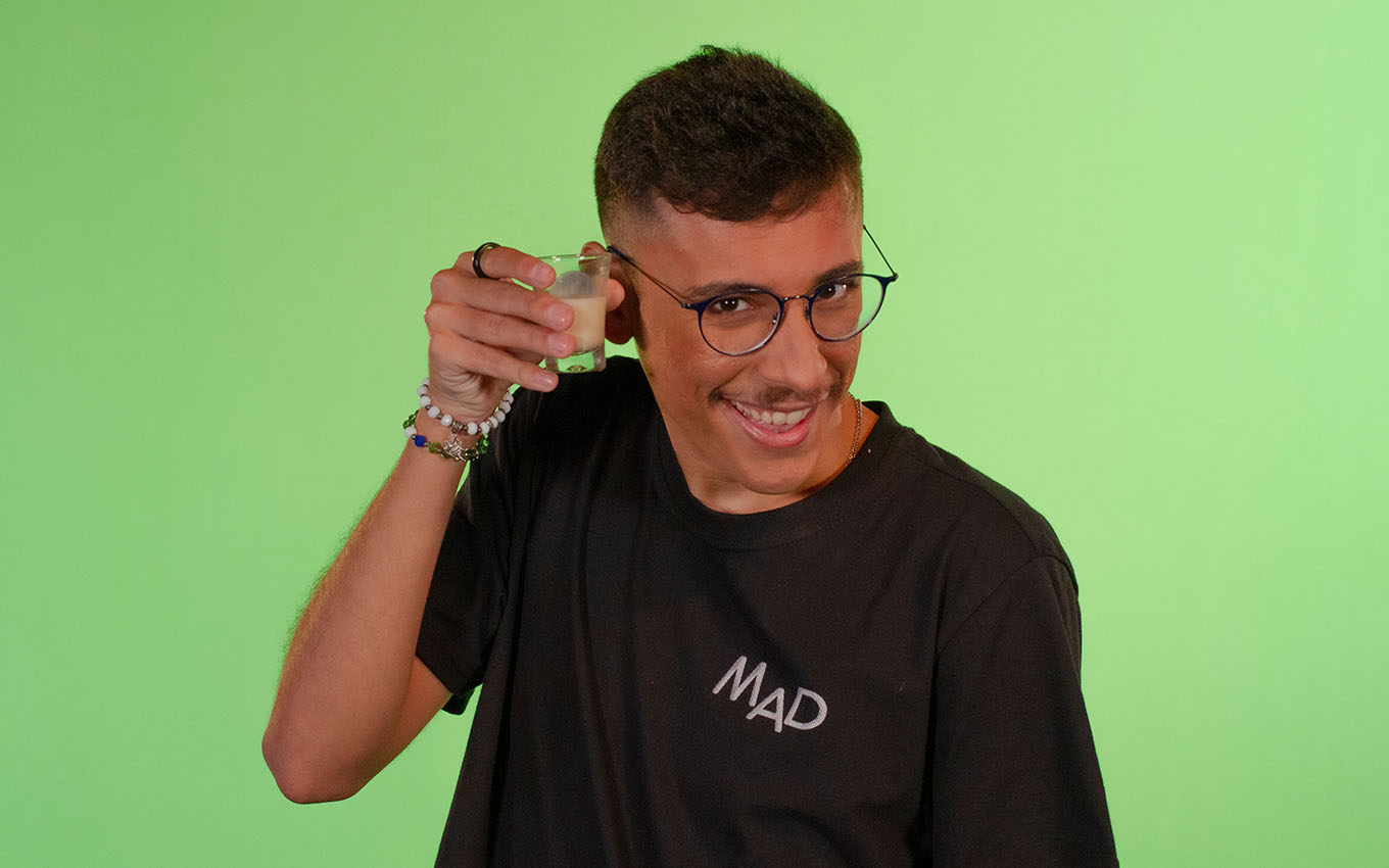 O comediante Matheus Mad segura um copo de batida diante de um fundo verde
