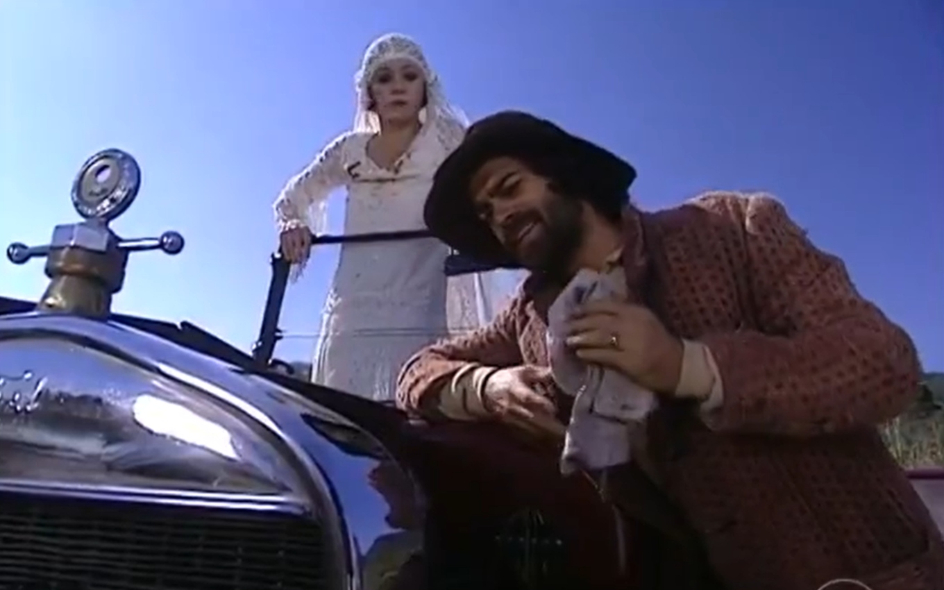 Adriana Esteves, a Catarina, está de pé no banco de um carro antigo sem teto, enquanto Eduardo Moscovis, o Petruchio, finge estar consertando o automóvel em cena de O Cravo e a Rosa