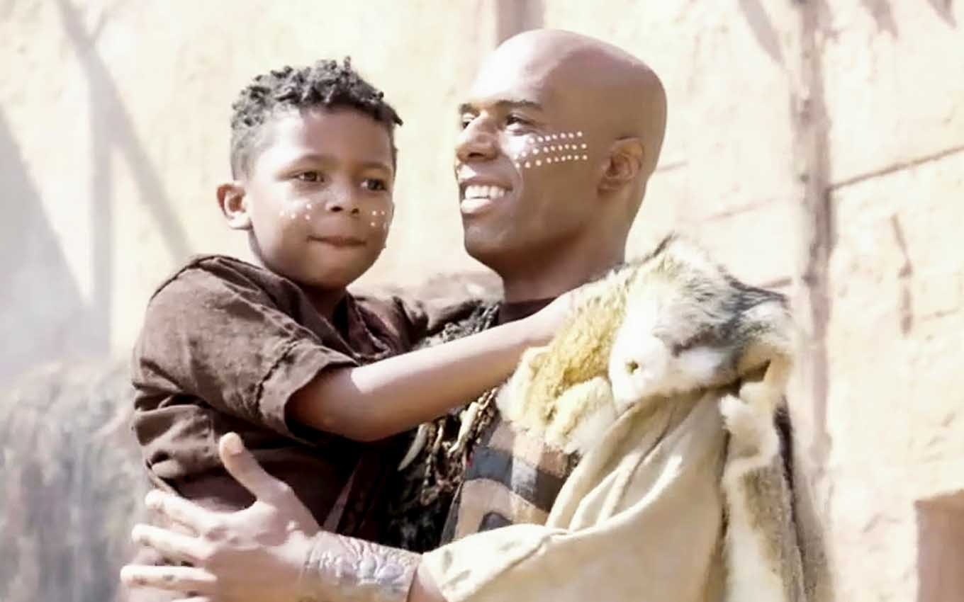 O ator Dudu de Oliveira como Abumani segura uma criança por volta de seis anos no colo em cena de Gênesis